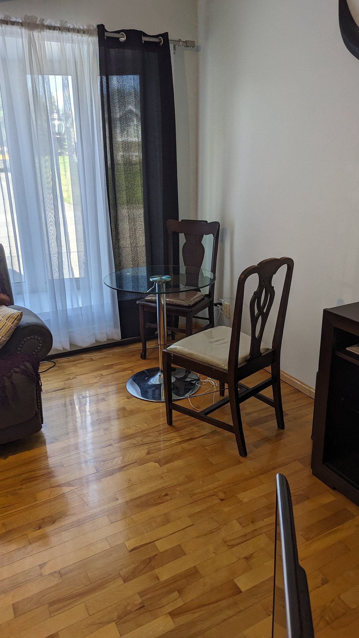 A small dining area features a round glass table accompanied by two wooden chairs with padded seats. Natural light filters through sheer curtains, illuminating the hardwood flooring and creating a bright and welcoming atmosphere.