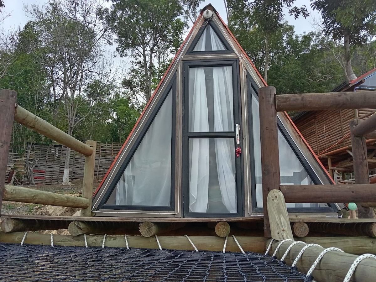 A triangular chalet is presented with large glass panels, allowing for ample natural light in the interior. It is elevated on wooden supports and surrounded by trees. A netted area in the foreground provides an additional recreational space.