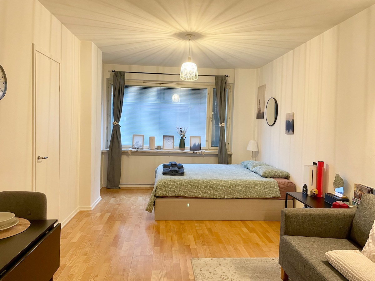 A spacious sleeping area is presented with a large bed and soft bedding. Natural light fills the room through a large window, adorned with curtain panels. A small table and a cozy seating area are visible, enhancing the inviting space.