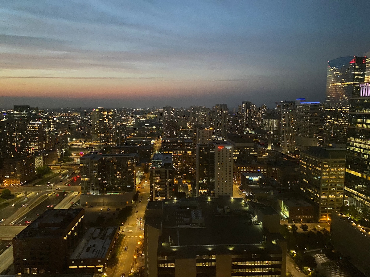 A panoramic view of the Chicago skyline at dusk showcases a blend of city lights and twilight hues. Tall buildings are observed against a gradient sky, transitioning from soft blue to warm orange. Streets below are lined with illuminated vehicles, indicating the city's vibrant life.
