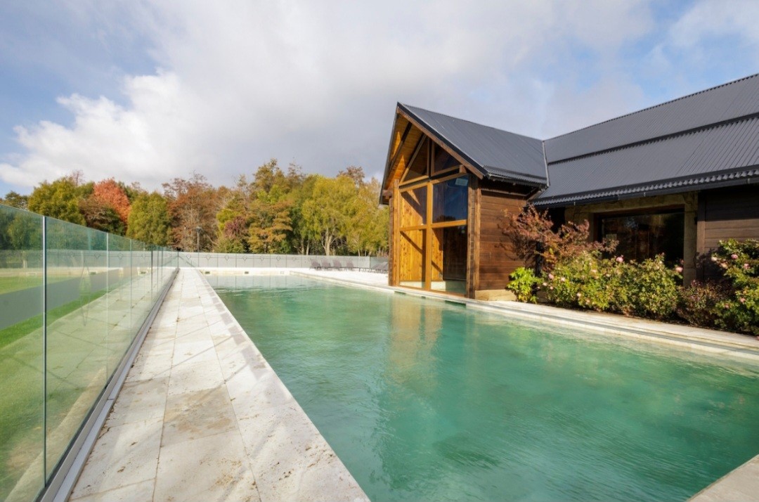 A serene swimming pool is surrounded by a glass fence, providing unobstructed views of the lush greenery beyond. The nearby wooden structure features large windows, blending with the natural surroundings. The pool area is designed for relaxation, showcasing a clean and inviting atmosphere.