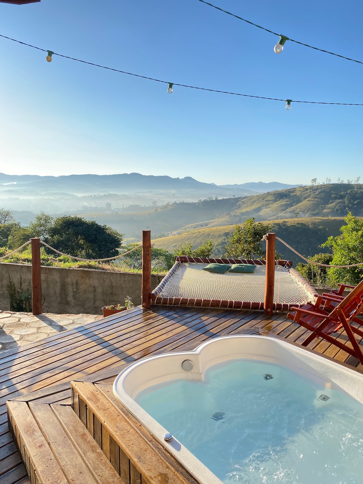 A wooden deck features a hot tub for two, surrounded by scenic views of rolling hills and mountains. A suspended hammock offers a relaxing spot, while string lights illuminate the space. Two red chairs are positioned near the hot tub, enhancing the outdoor experience.
