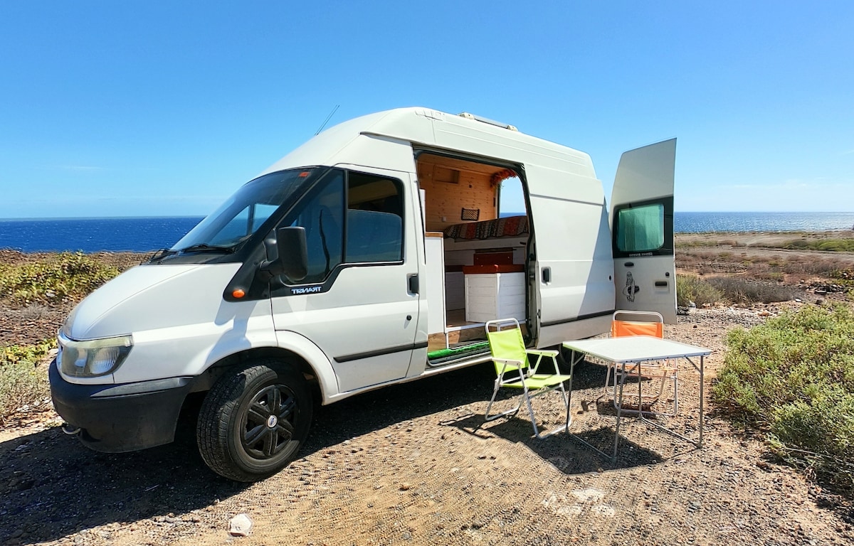 A camper van is parked on rocky terrain near the coast, with ocean views in the background. The door is open, revealing a compact interior setup. A small outdoor table and two colorful chairs are positioned beside the van.