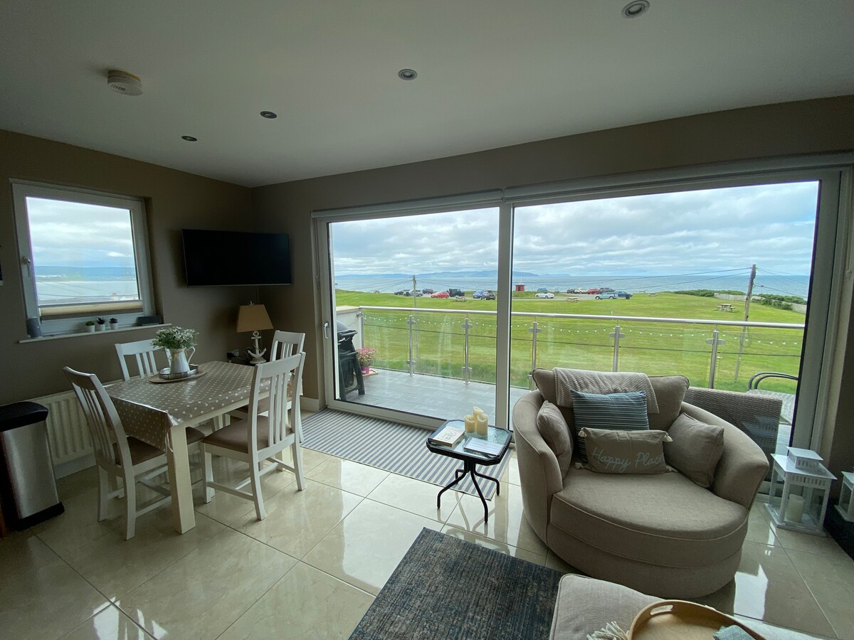The open-plan living area features large sliding glass doors that provide panoramic views of the green landscape and distant sea. A cozy armchair is paired with a small table, while a dining table with chairs is positioned nearby. Natural light floods the space, enhancing the modern decor.
