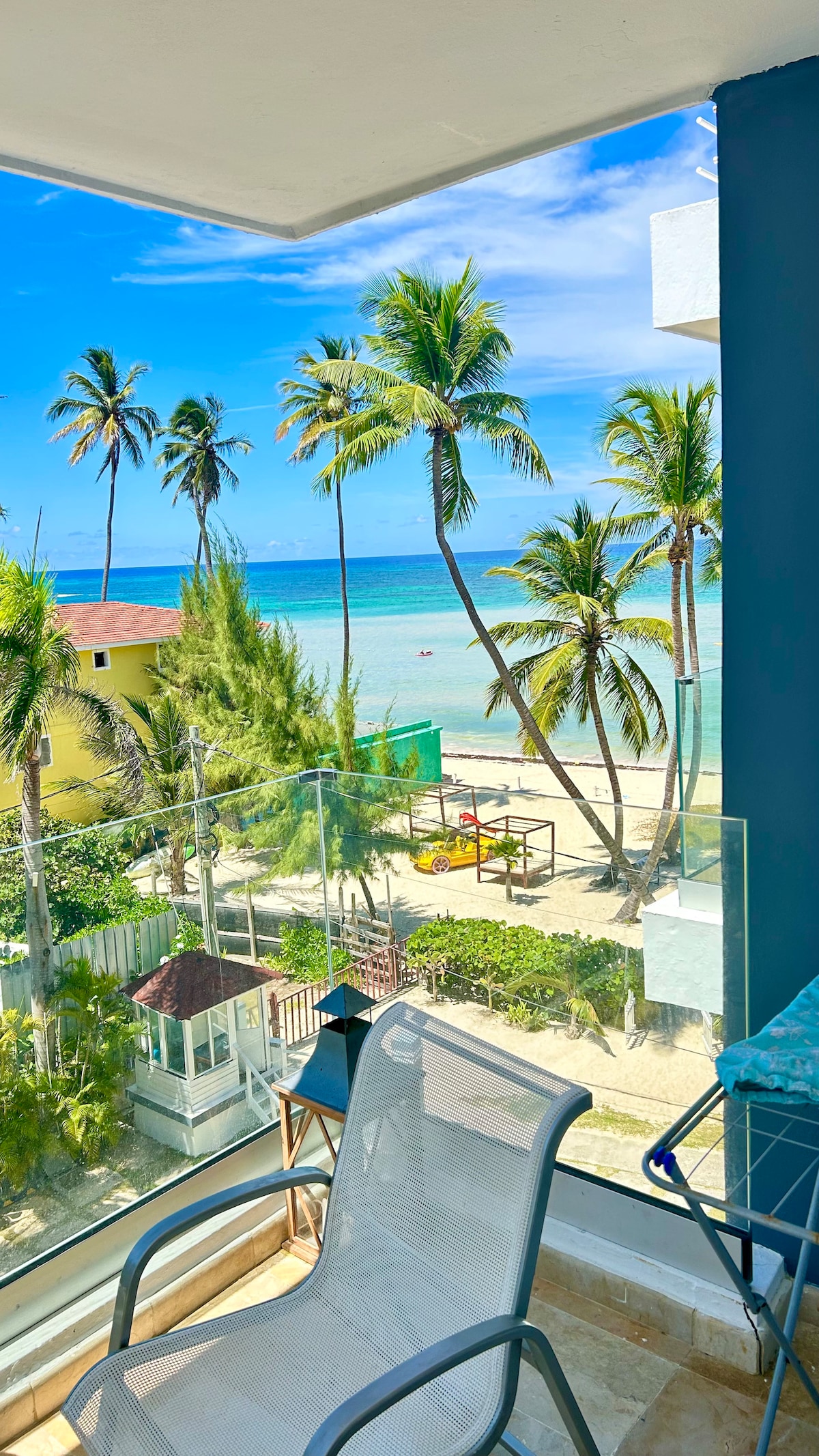 A view from the fourth floor features palm trees swaying against a bright blue sky, overlooking a serene beach. The glass balcony provides an unobstructed perspective of the turquoise water, where a few small boats can be seen in the distance, adding to the coastal ambiance.