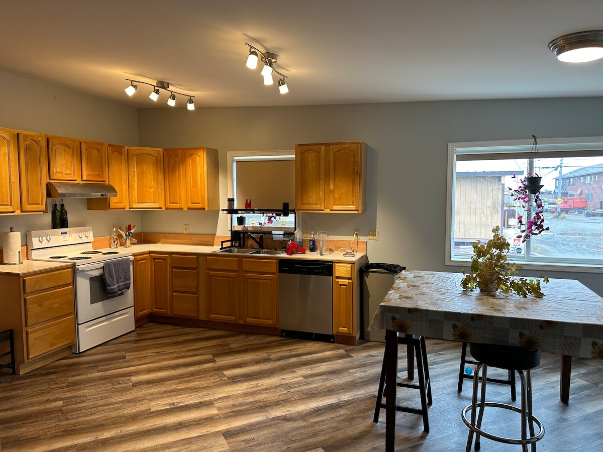 The kitchen features wooden cabinetry and modern appliances, with a window overlooking an exterior view. A central table is accompanied by high stools, providing a casual dining space. Natural light illuminates the area, highlighting the wood flooring throughout.