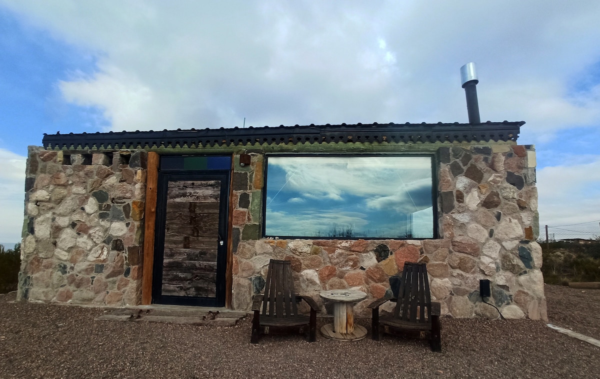 A stone cabin with a minimalist design features a large window reflecting the sky. Two wooden chairs are positioned in front of the entrance, accompanied by a small circular table. The exterior showcases natural stonework, blending into the surrounding landscape.