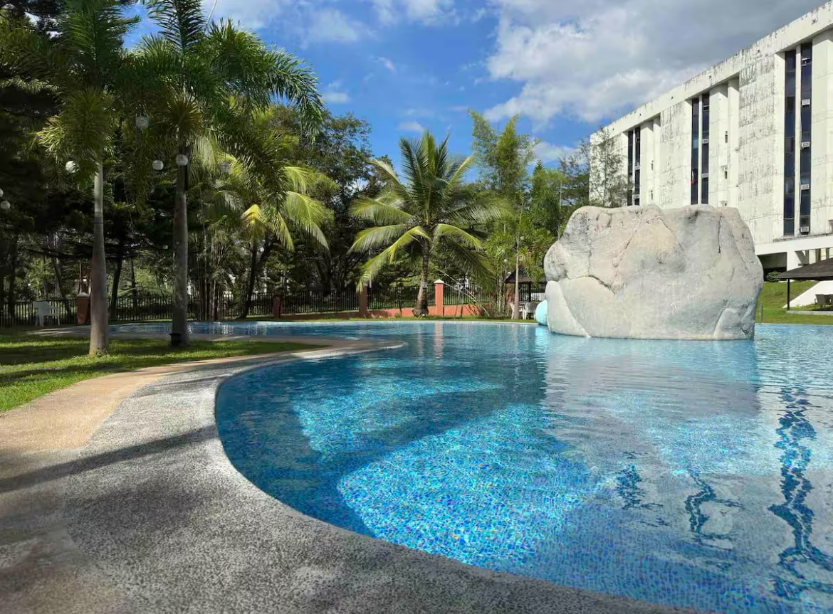 A tranquil pool area is set among lush greenery, with palm trees providing shade. A large rock formation is positioned in the water, surrounded by a gently curved edge of the pool. Clear blue water reflects the sky, creating a serene atmosphere.