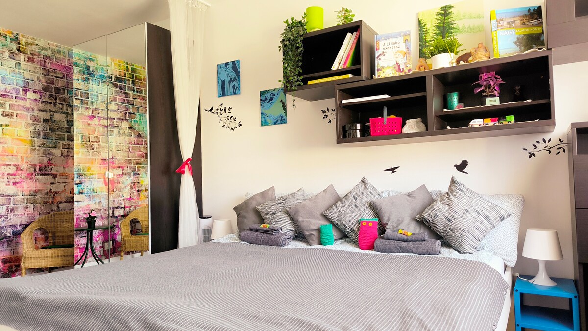 A spacious sleeping area features a large bed adorned with several decorative pillows. Behind the bed, a vibrant mural adds color to the wall, while shelves display books and plants. A comfortable chair is visible in the corner, enhancing the inviting atmosphere.