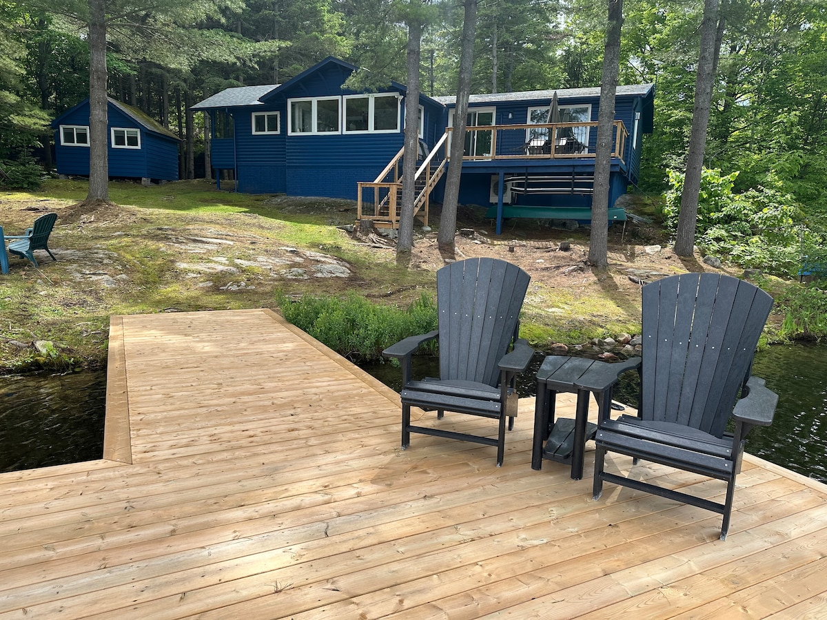 A wooden dock extends over calm waters, featuring two adirondack chairs and a small side table inviting relaxation. In the background, a blue cottage is nestled among trees, showcasing large windows and a spacious deck, harmonizing with the serene woodland setting.