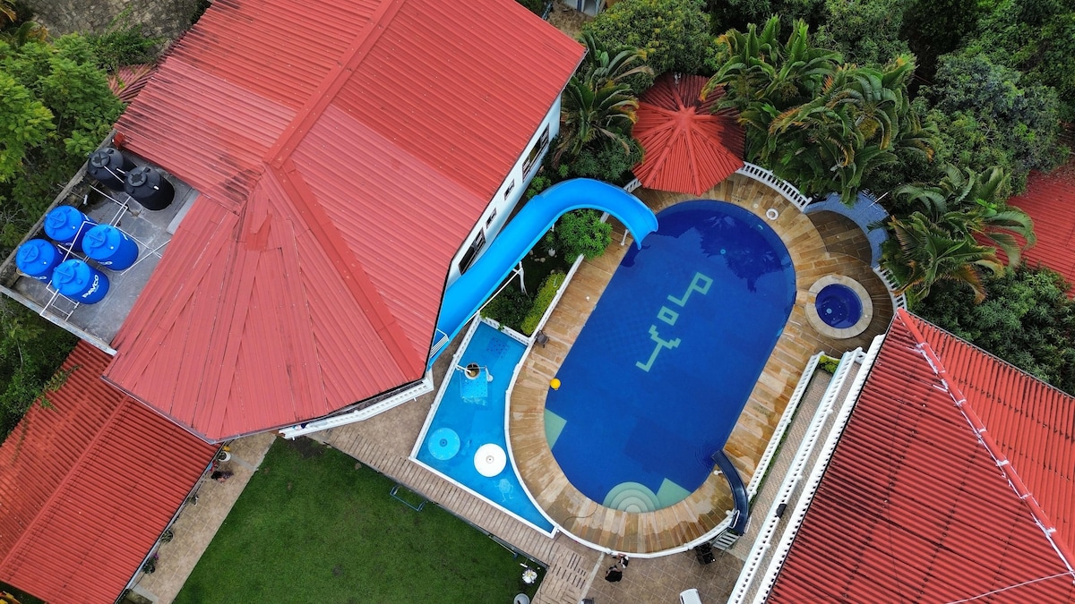 The outdoor area features a large swimming pool with a blue waterslide, surrounded by a wooden deck. A separate section with seating and a smaller circular pool can be seen. Lush greenery and palm trees provide natural landscaping around the space.