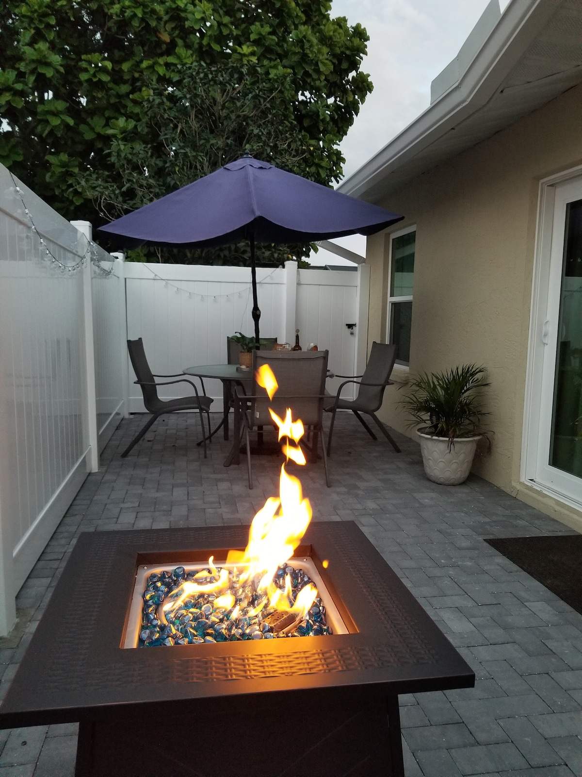 A private patio area is featured, furnished with a table and four chairs beneath a purple umbrella. A fire pit with colorful glass stones is lit, providing a cozy ambiance. The setting is surrounded by a white fence and lush greenery.
