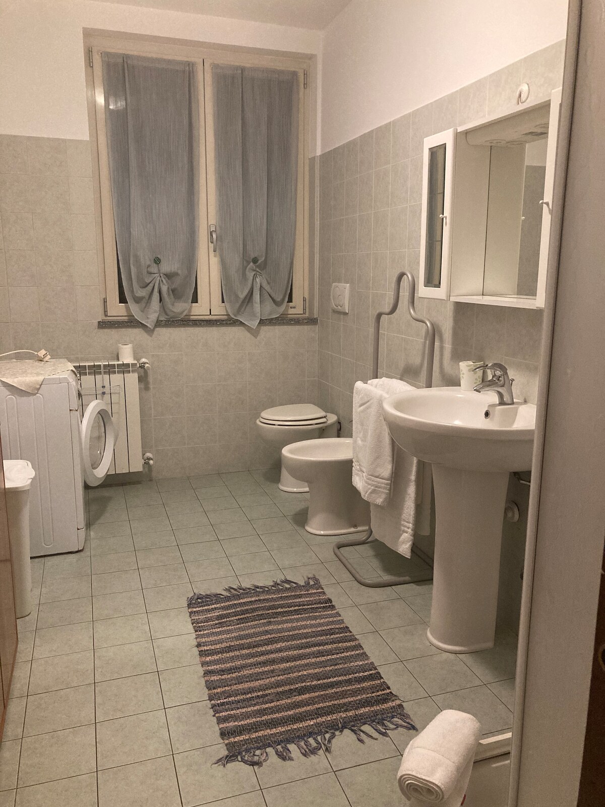 The bathroom features light-colored tiles and is equipped with a washing machine, a toilet, a sink with a mirror, and a towel rack. Two windows with light drapes provide natural light. A small striped rug is placed on the floor, adding a touch of comfort.