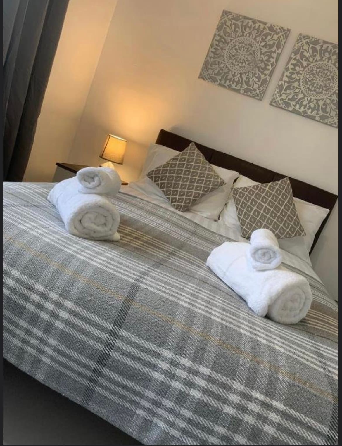 A well-styled bedroom features a spacious bed adorned with a patterned blanket. Two rolled towels are placed on the bed, and soft lighting from a bedside lamp creates a cozy ambiance. Decorative wall art adds a touch of character.
