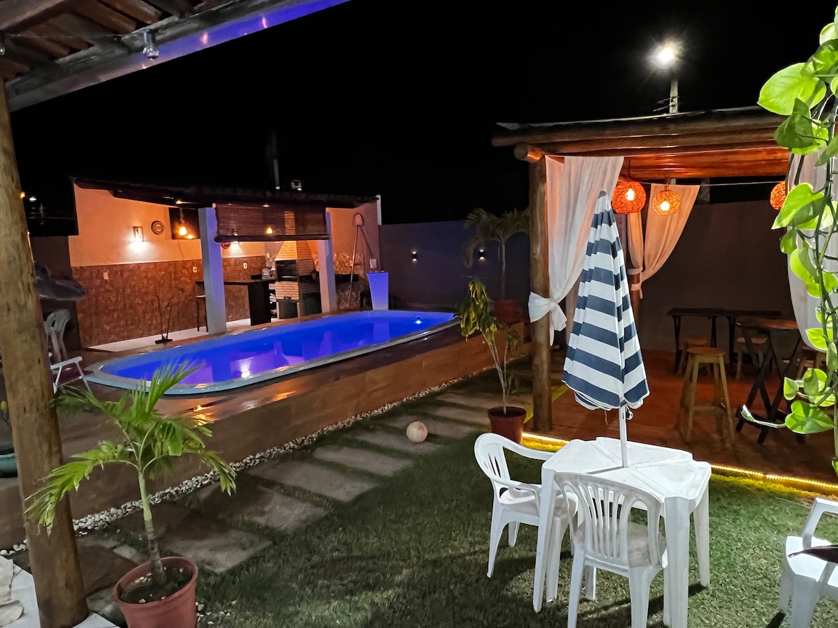 A well-lit outdoor area showcases a vibrant pool featuring illuminated water. A shaded seating area with a table and chairs invites relaxation, while decorative plants create a welcoming atmosphere. The space is further enhanced by string lights and a pergola, providing a cozy feel at night.