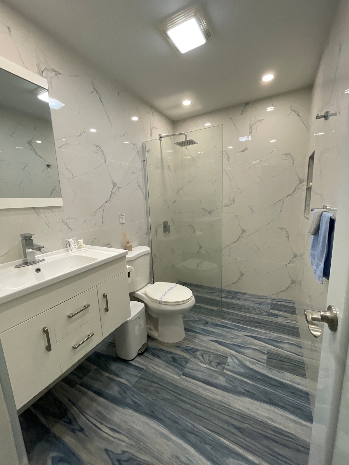 The bathroom features sleek marble walls with a glass shower enclosure. A modern vanity with a spacious countertop and a large mirror is visible. A toilet and a small waste bin are also present, accompanied by a towel hanging on the wall.