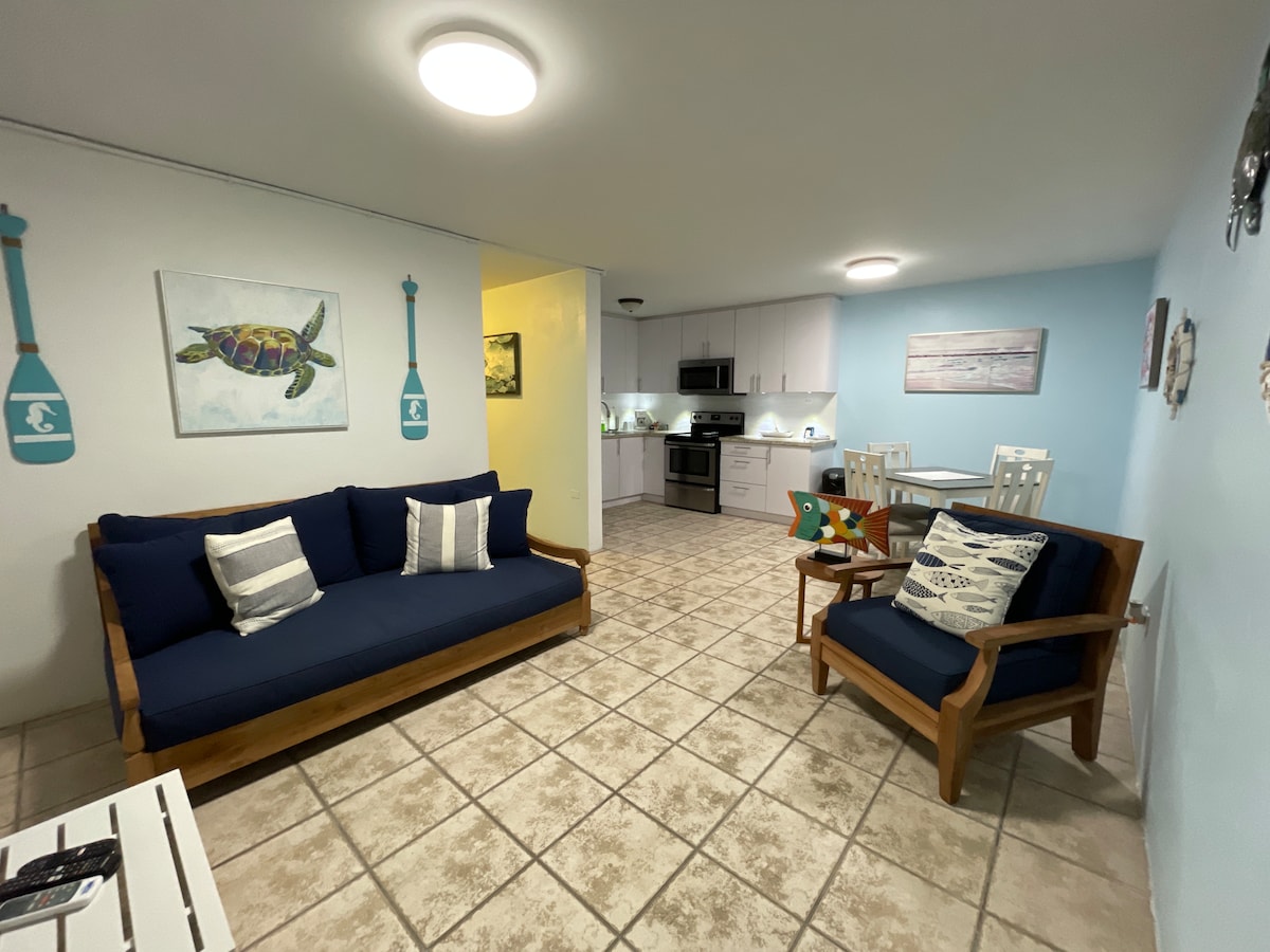A comfortable living area features a dark blue sofa and a wooden armchair, complemented by decorative cushions. Light tiles cover the floor, and soft blue walls enhance the inviting atmosphere. Two paintings of a turtle and a beach scene are displayed on the wall.