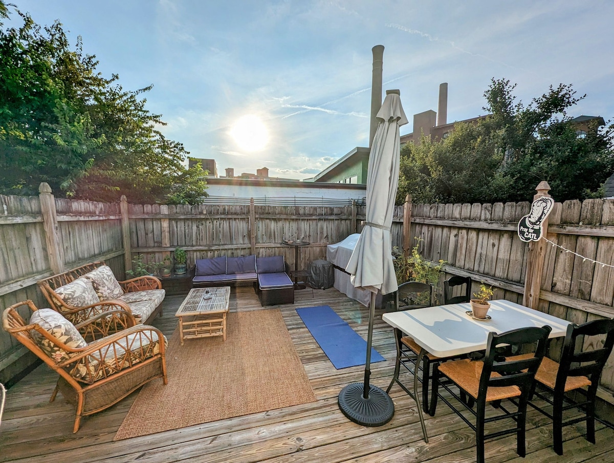 An outdoor deck area features a mix of seating options, including rattan chairs and a rectangular table with four black chairs. A yoga mat is placed on the wooden floor. Sunlight sets in the background, casting a warm glow over the space.