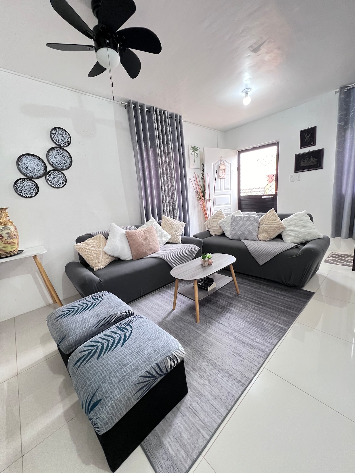 A well-lit living area features a grey sofa adorned with several decorative cushions. A small round table is positioned in front, with a soft grey rug underneath. A fan hangs from the ceiling, and light curtains frame the window, allowing natural light to enter.