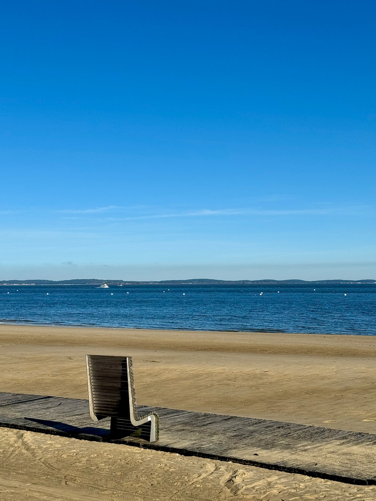 A serene beach view is captured under a clear blue sky, featuring soft sandy shores and calm waters. Gentle waves lap the beach, with distant hills visible on the horizon, creating a peaceful coastal landscape.
