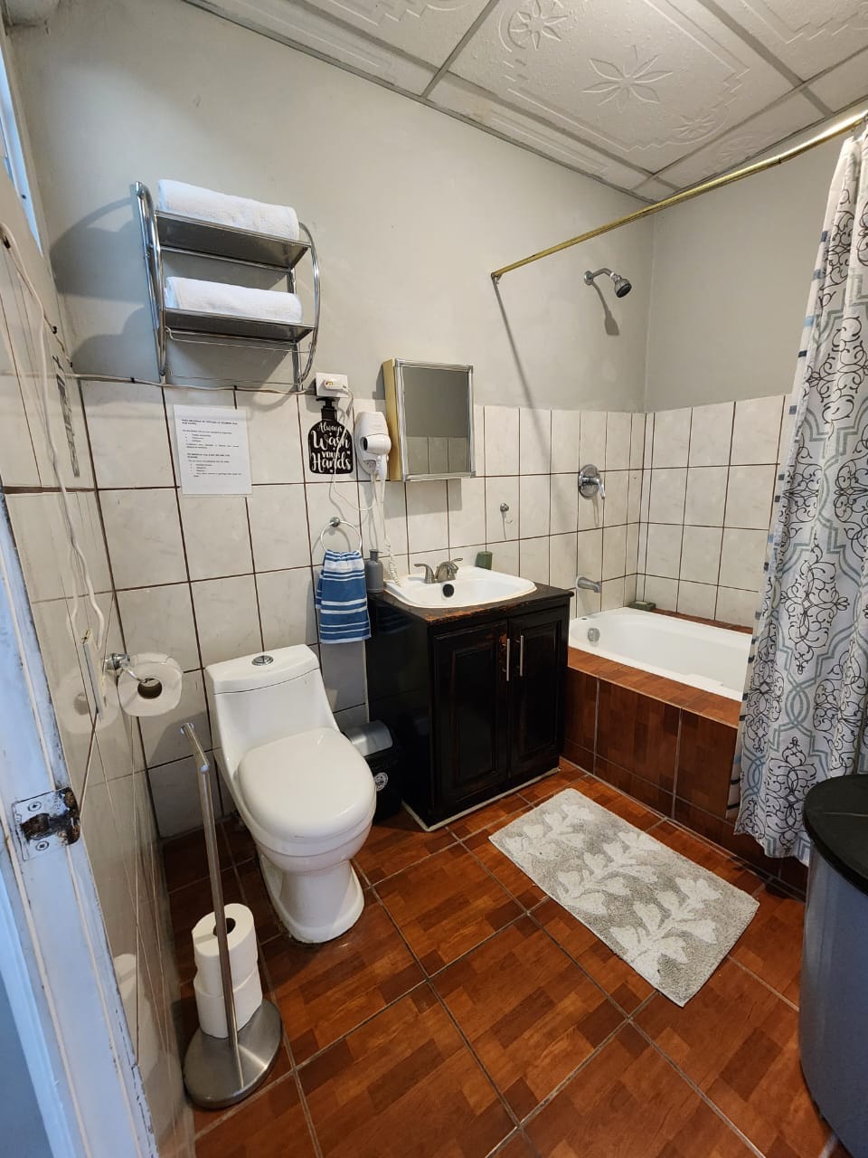 The bathroom features a combination of tiled surfaces and wood flooring. A toilet and a dark wooden sink cabinet are positioned beside a bathtub, which is enclosed by a shower curtain. Clean towels and toiletries are neatly arranged on a shelf, with a small rug on the floor.