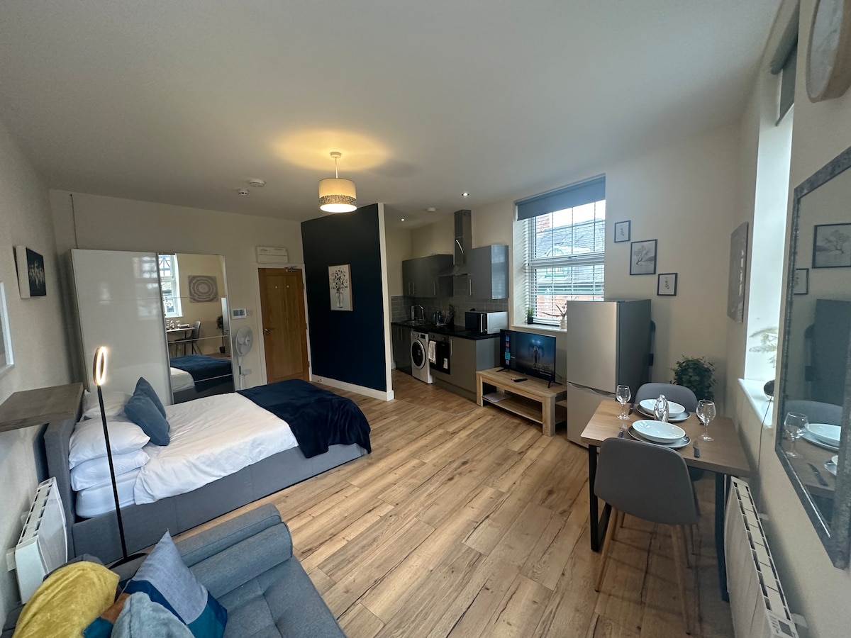An open-plan studio features a comfortable double bed and a grey sofa, complemented by wooden floors. A compact dining table for two and a well-equipped kitchen, including a washer/dryer and refrigerator, are visible. Natural light enters through the window, enhancing the space.