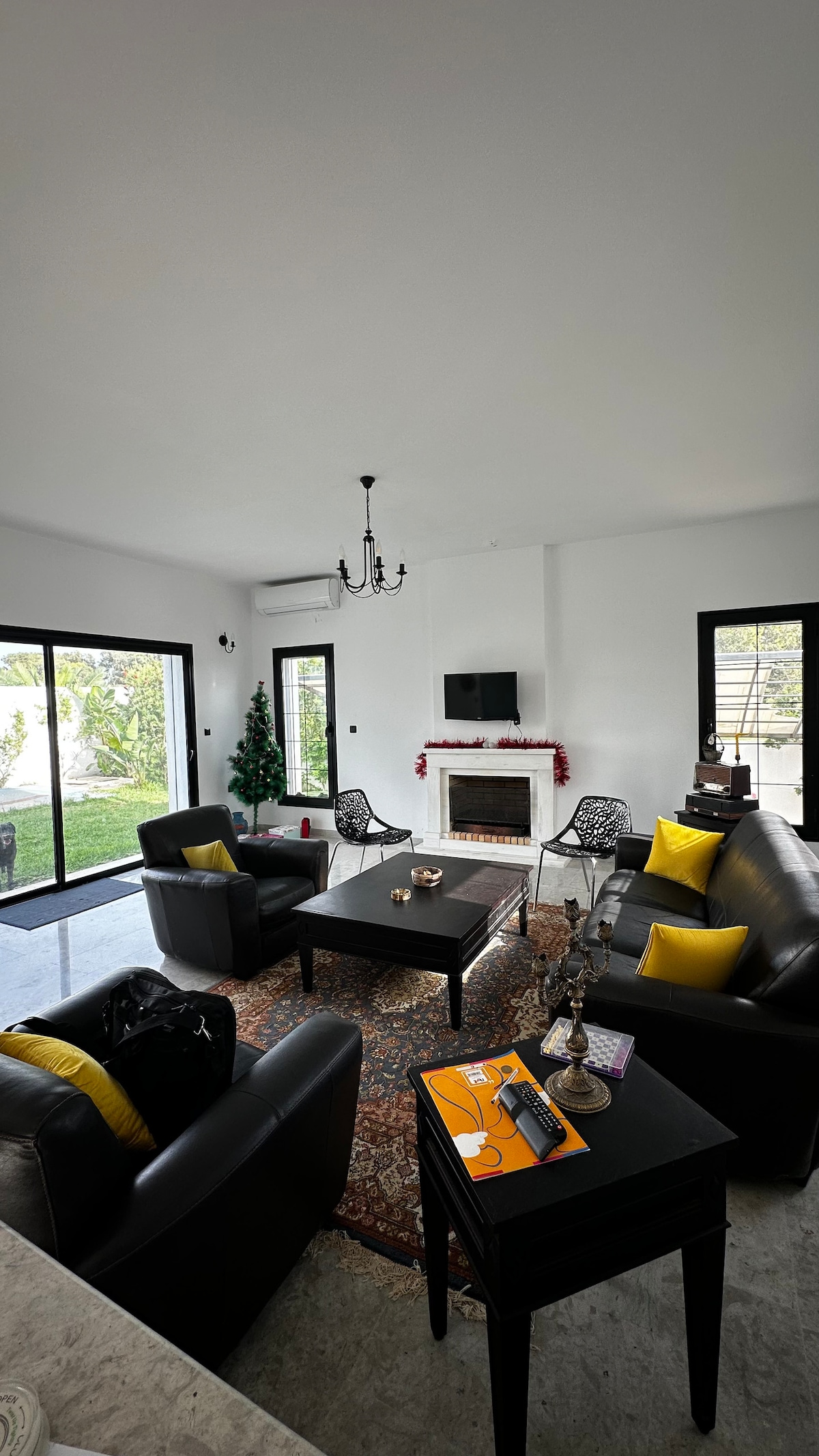 A spacious living area features dark leather sofas paired with yellow accent pillows, centered around a coffee table on a patterned rug. Large windows provide natural light and views of the outdoors. A flat-screen TV is mounted on the white wall, and a decorative fireplace adds warmth.