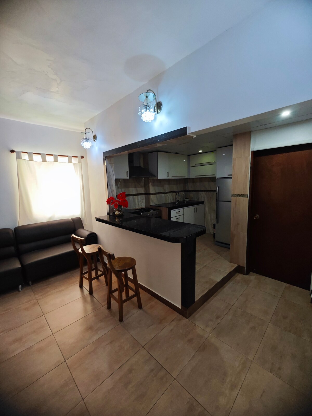 A modern kitchen with a breakfast bar is shown, featuring two wooden stools. The space is illuminated by pendant lights, and white cabinetry complements the dark countertops. A cozy seating area with a dark sofa is visible, with natural light filtering through a window adorned with light curtains.