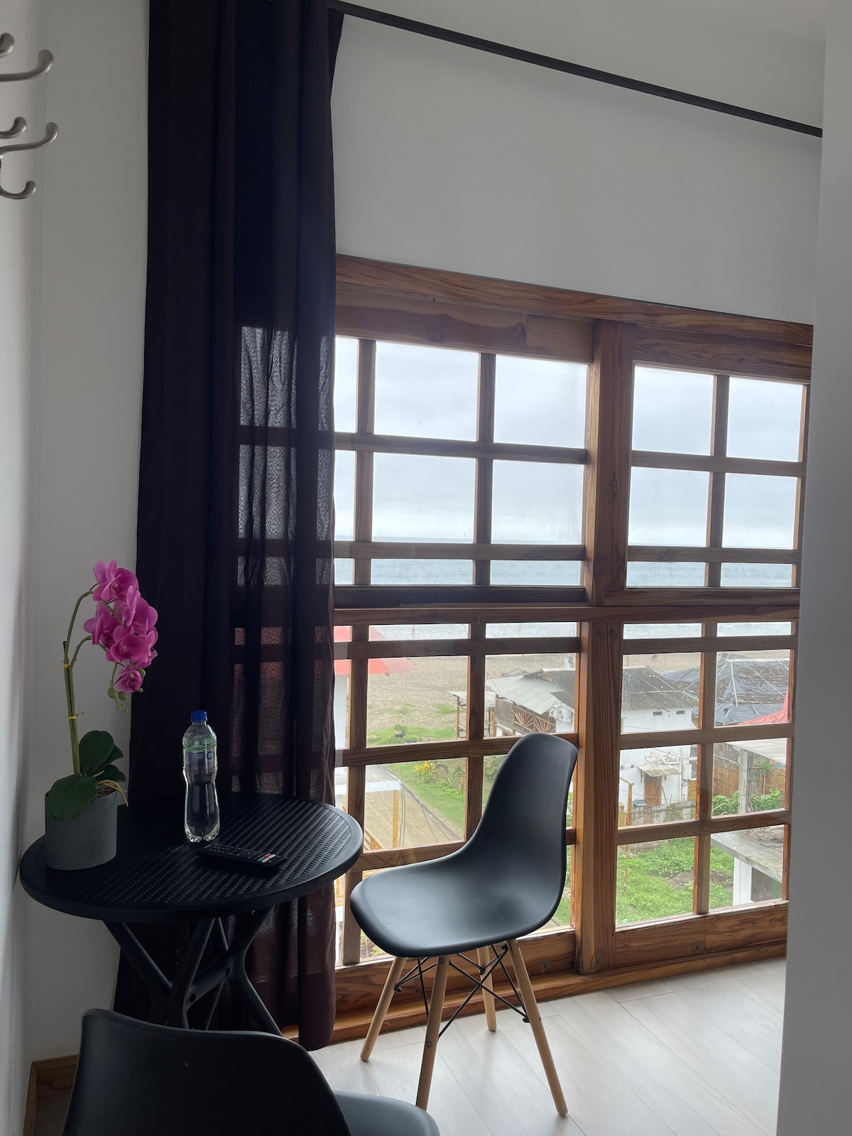 A light-filled corner features a small round table and two black chairs, overlooking a window with a grid pattern. Dark curtains frame the window, complementing a nearby potted plant with a vibrant flower and a bottle of water.