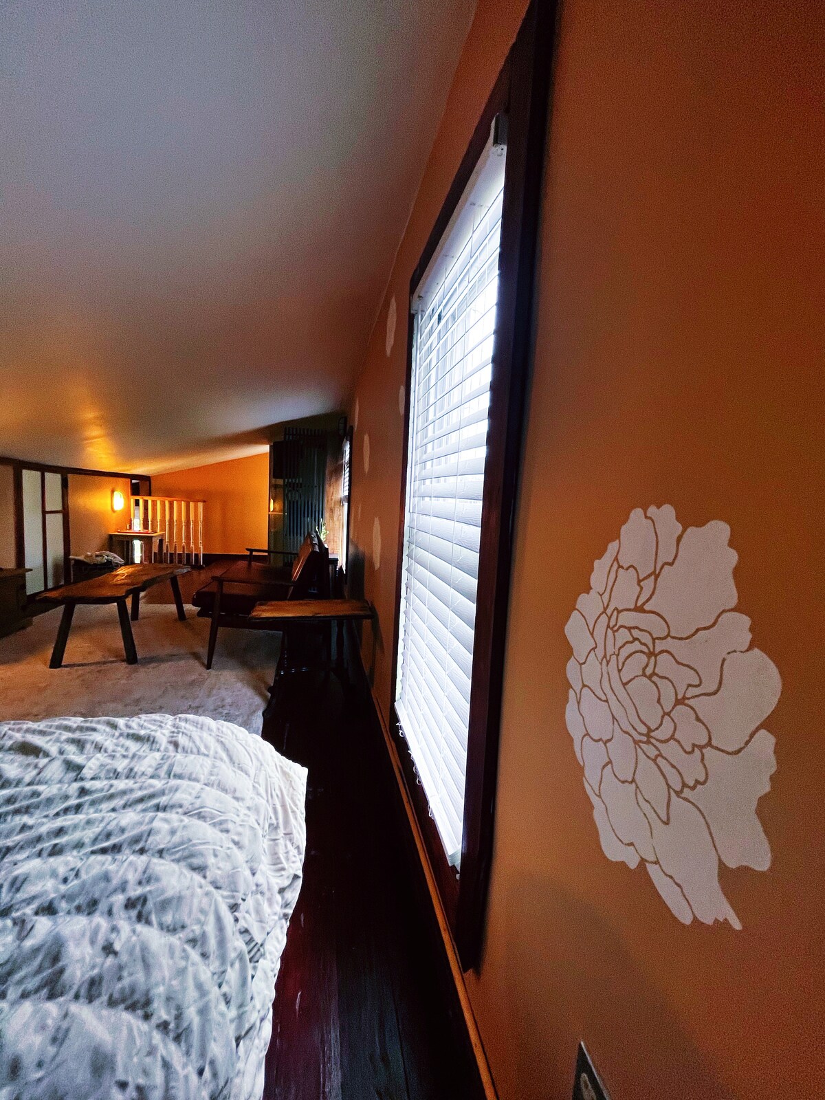 An open sleeping loft features a low slanted ceiling and is adorned with warm earthy tones. A white floral design is painted on the wall beside a window with blinds, allowing soft light to brighten the space. A cozy bed and small seating area are visible.