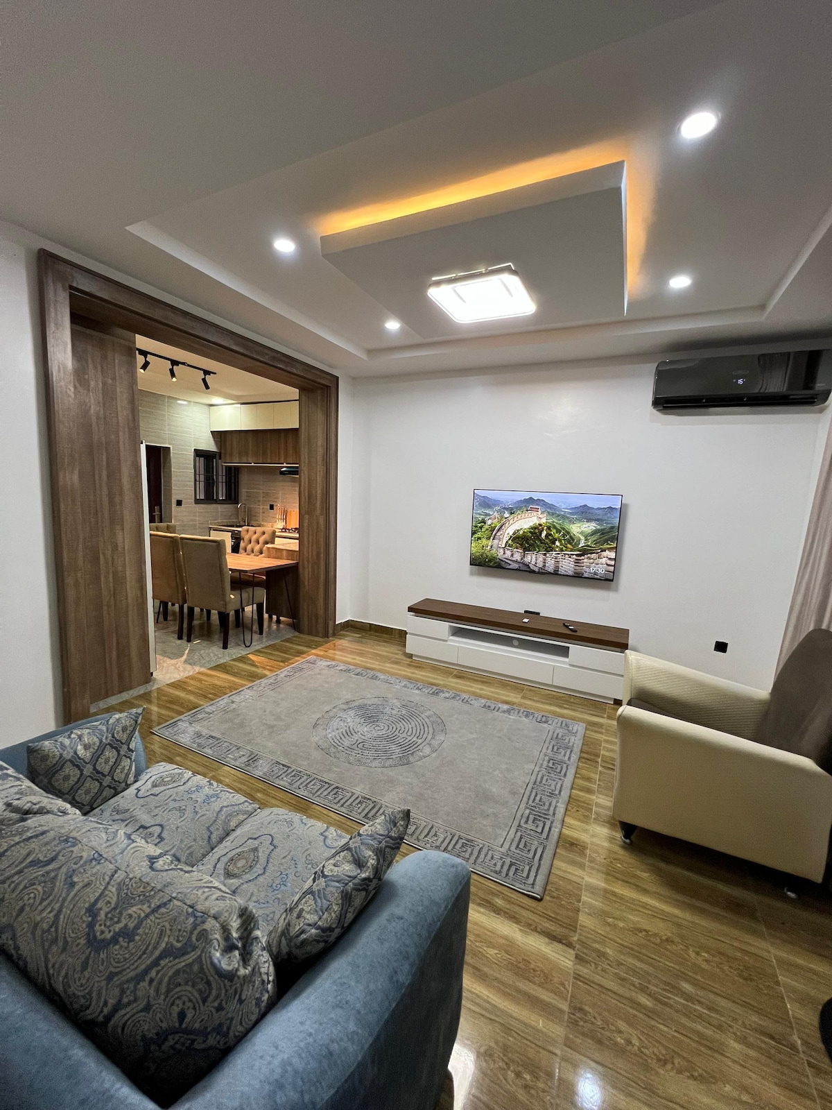 A contemporary living area is highlighted by a large area rug and comfortable seating options, including a blue sofa and a beige chair. A flat-screen TV is mounted on the wall, and a dining area is visible in the background, showcasing a modern kitchen space.