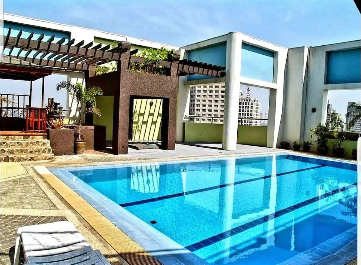 A spacious rooftop pool is surrounded by modern architecture, featuring a sunlit deck. Lounge chairs are positioned alongside the clear water. A shaded pergola with greenery enhances the area, providing a relaxing space for guests to unwind while enjoying views of the city.