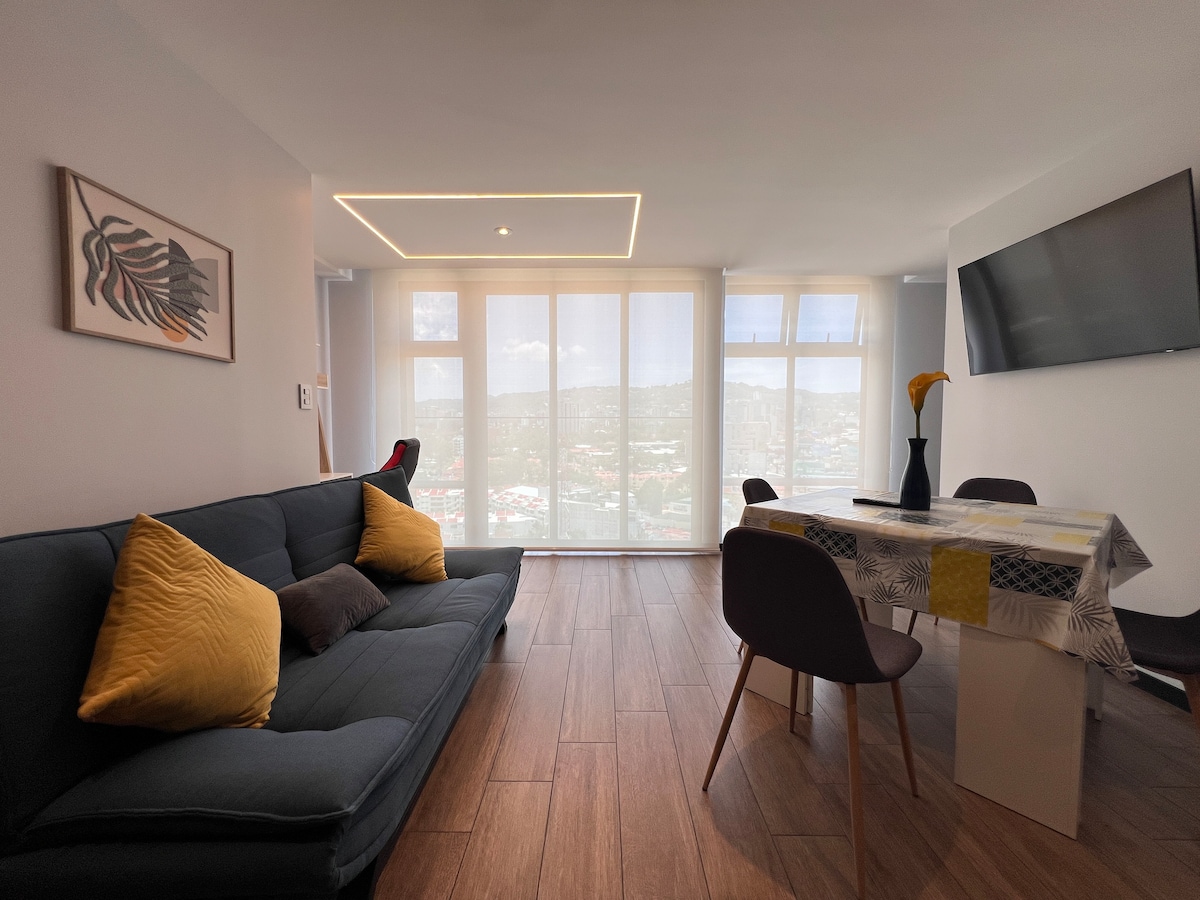 A modern living area features a dark blue sofa with bright yellow cushions, paired with a dining table set for four. Large windows flood the space with natural light and offer expansive city views. A wall-mounted television and a decorative vase add to the decor.