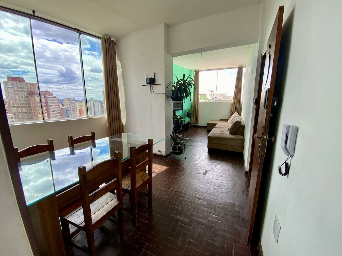 A bright living area features large windows allowing natural light to fill the space. A comfortable sofa is arranged beside a television, while a dining table with wooden chairs provides a setting for meals. Indoor plants add a touch of greenery to the room.