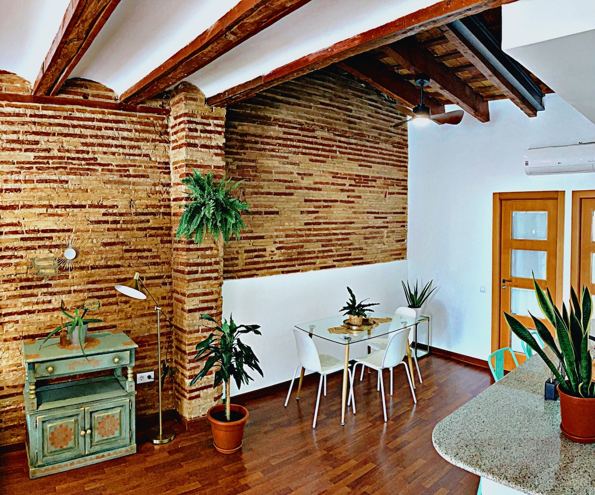 A dining area features a glass table surrounded by white chairs, complemented by lush indoor plants. The space is defined by exposed brick walls and wooden beams overhead, creating a unique architectural style. A decorative desk is positioned nearby, enhancing the inviting atmosphere.