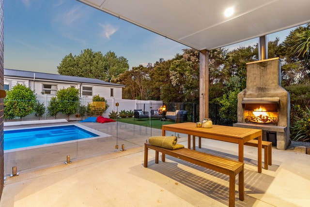 An inviting outdoor space features a swimming pool surrounded by greenery. A wooden dining table with benches is positioned nearby, complemented by an outdoor fireplace. Cozy seating options are visible, creating a serene atmosphere for relaxation or gatherings.