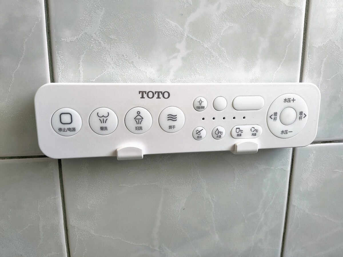 A smart bidet toilet seat control panel is mounted on a tiled wall. The panel is white with various buttons labeled with icons for different features, providing convenience and comfort for users.