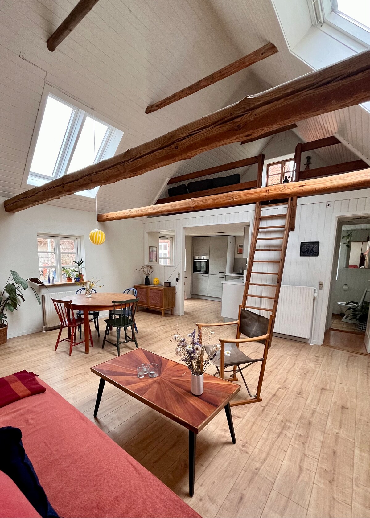 An open living area features high ceilings with exposed beams and large windows allowing natural light. A cozy seating area is arranged with a sofa and coffee table. A dining table and chairs sit nearby, and a wooden ladder leads to an upper loft space.
