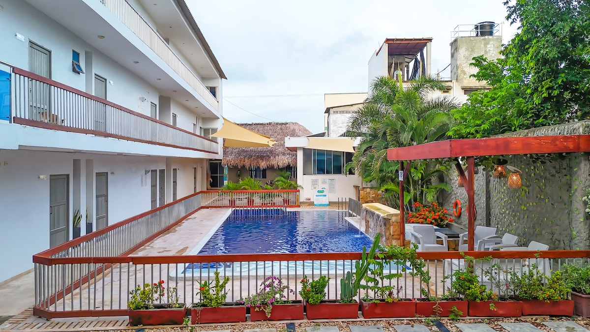 A swimming pool is surrounded by a wooden deck and tropical plants, with lounge chairs situated nearby. The two-story building features a row of doors leading to individual rooms, and a shaded area with thatched roofing is visible in the background.