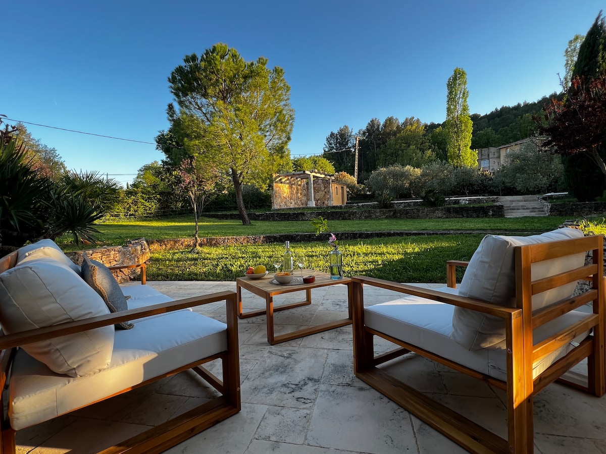 An outdoor seating area is presented, featuring two comfortable chairs and a low table. The view opens onto a lush garden, with trees and greenery visible in the background. Natural stone flooring adds to the serene atmosphere of the space.