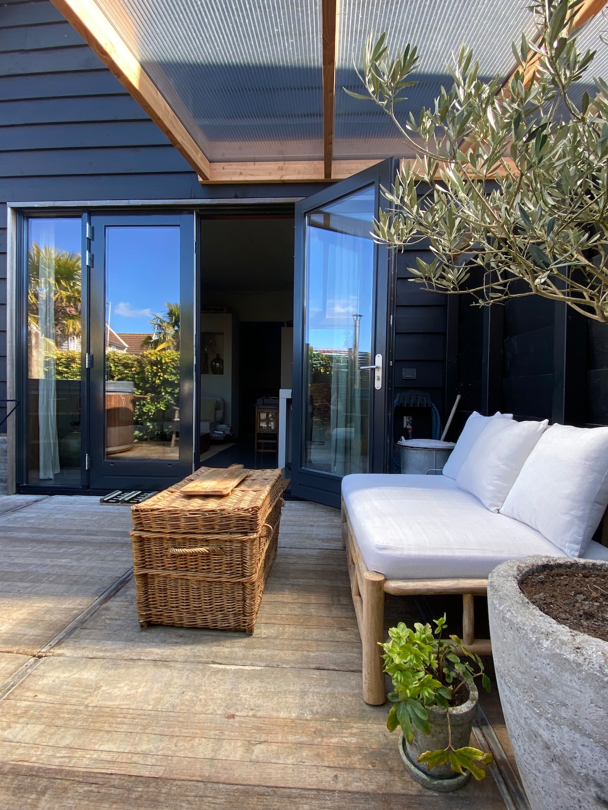 A private terrace features a cozy seating area with a white cushion-covered bench and a wicker coffee table. Glass doors open to the interior, allowing natural light to illuminate the space. Green plants and an olive tree add a touch of nature to the relaxed setting.