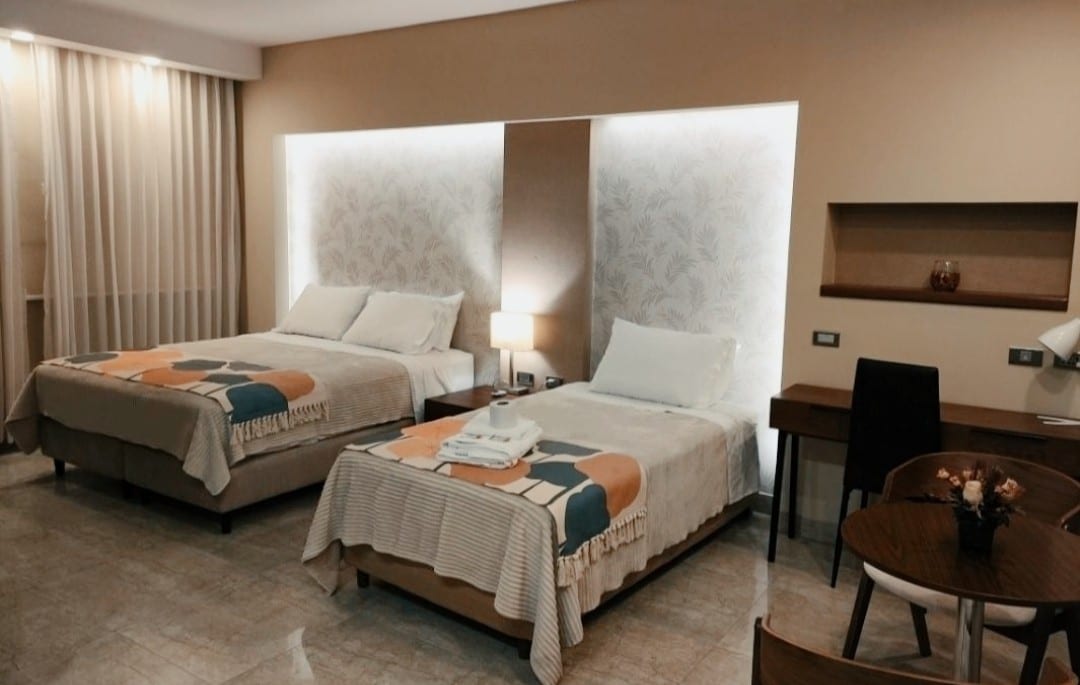 The room features two spacious beds, each adorned with soft linens and decorative throws. A soft light emanates from behind the headboards, highlighting the patterned accent wall. A small desk and a dining table are present, accompanied by a chair, creating a functional and welcoming space.
