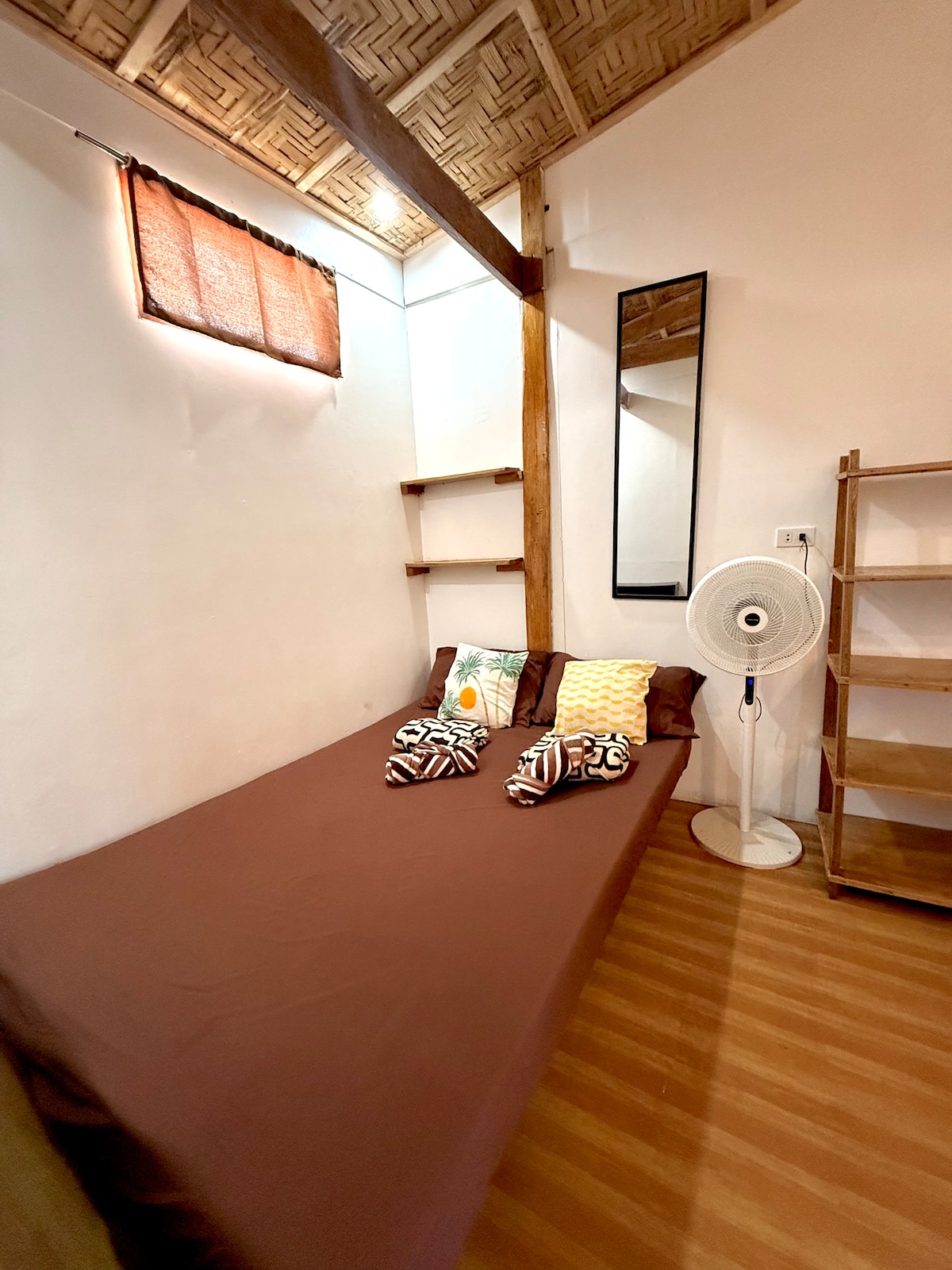 A cozy sleeping area features a spacious bed with a brown bedspread, complemented by two patterned pillows. A standing fan is positioned nearby, and a tall shelf offers additional storage. Natural light enters through a window with wooden blinds, enhancing the warm wooden flooring.
