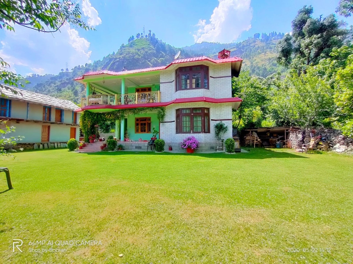 A two-story house is surrounded by a lush green lawn, featuring a spacious balcony with decorative potted plants. The exterior displays a combination of white and light red colors, with multiple windows reflecting the natural light. Tall trees and mountains frame the background.