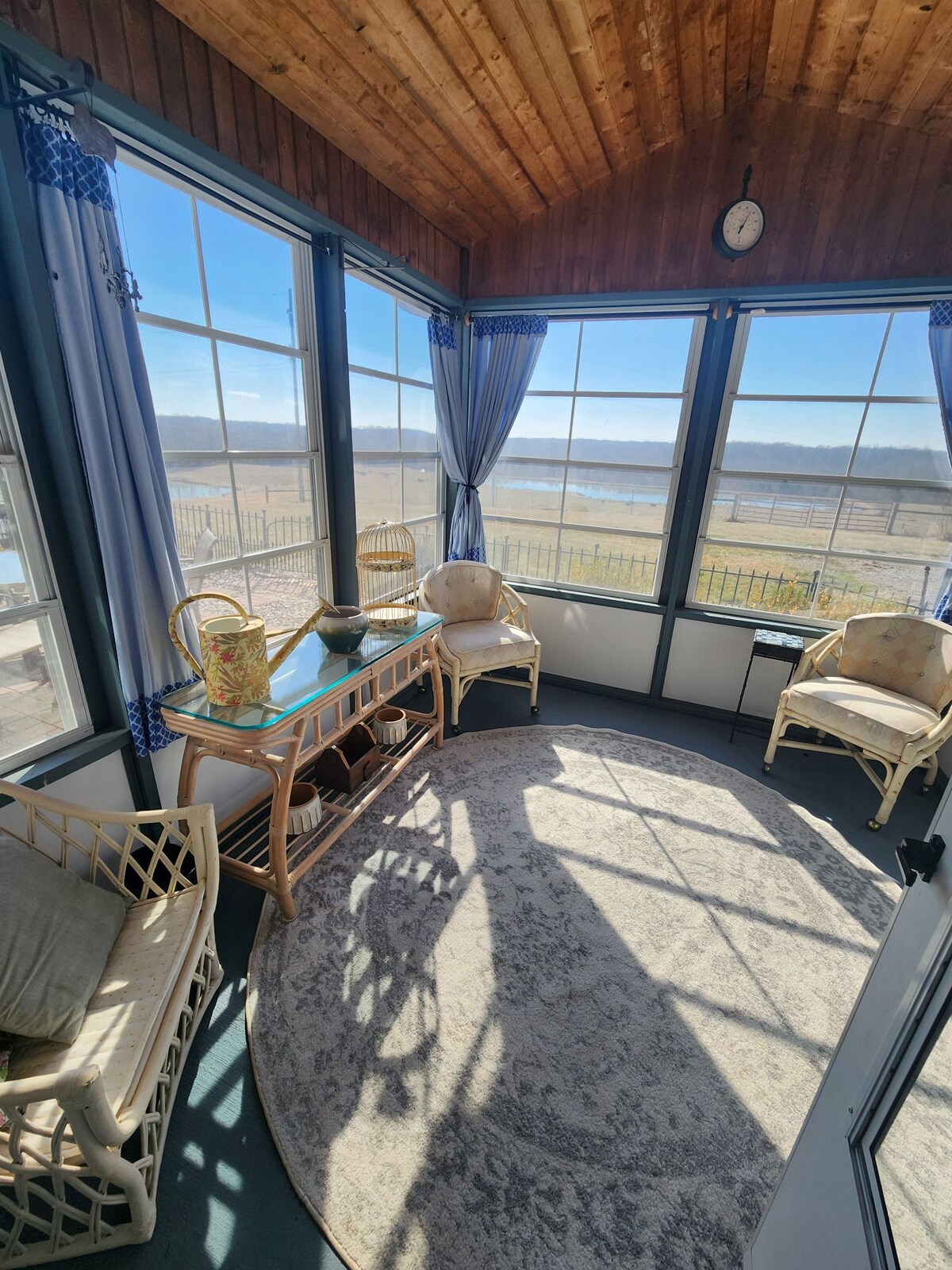 A sunlit three-season room features large windows offering expansive views of the countryside. A round rug is placed on a dark floor, surrounded by two light-colored chairs and a small table. Decorative curtains frame the windows, enhancing the inviting atmosphere.