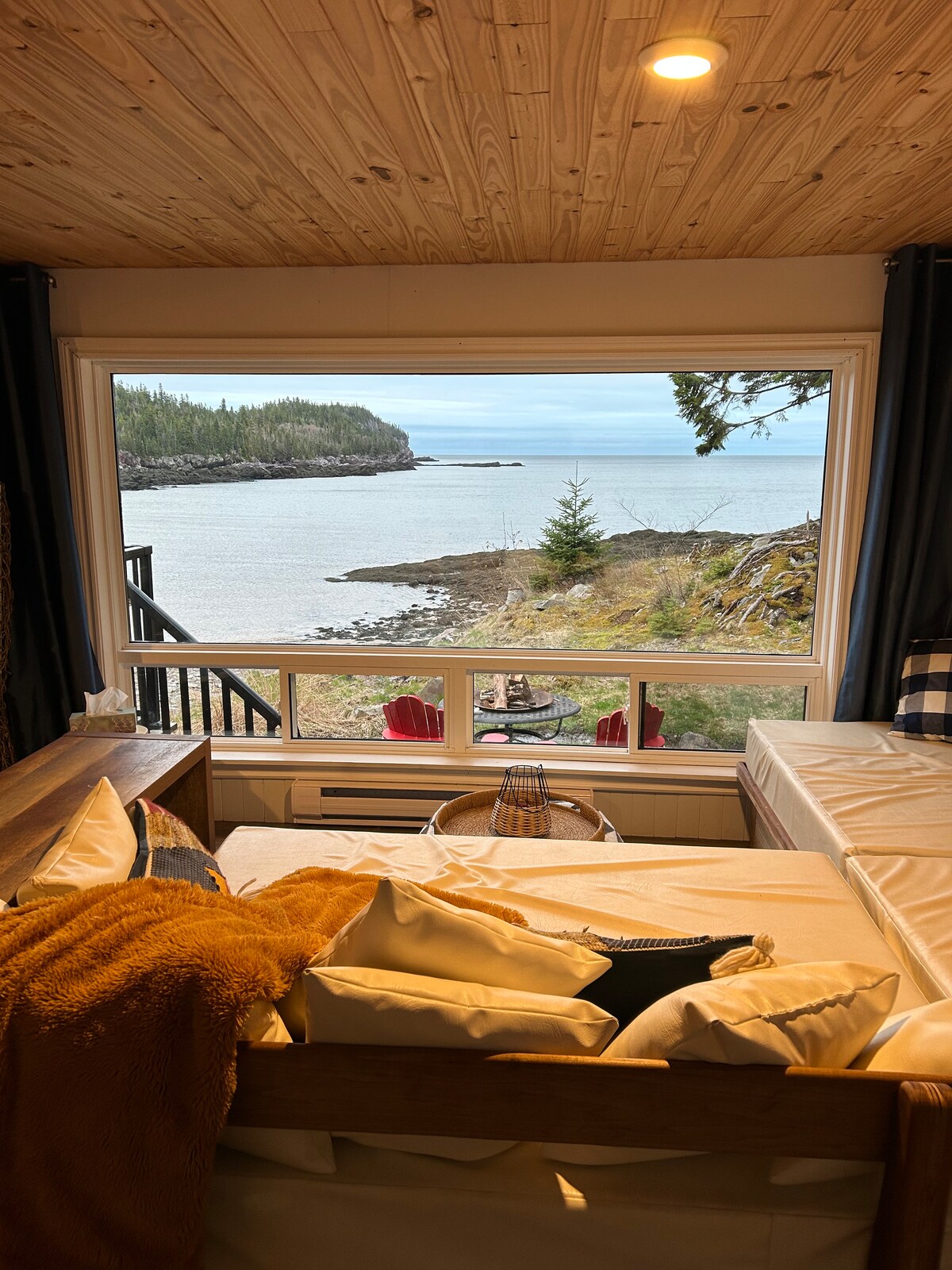Large windows frame a serene view of the bay, where calm waters meet rocky shores. Soft seating with neutral cushions invites relaxation, while warm textiles add comfort. The arrangement creates a cozy nook, ideal for enjoying the surrounding natural beauty.