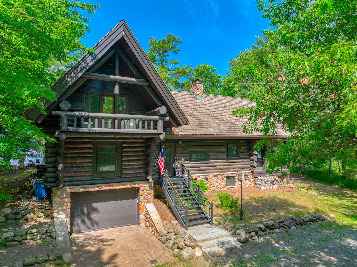 A spacious log cabin is nestled among lush greenery, featuring a stone-accented exterior and a welcoming front entrance. The building includes multiple gabled roofs and large windows, allowing natural light to flood the interior. A flag enhances the inviting ambiance of the home.