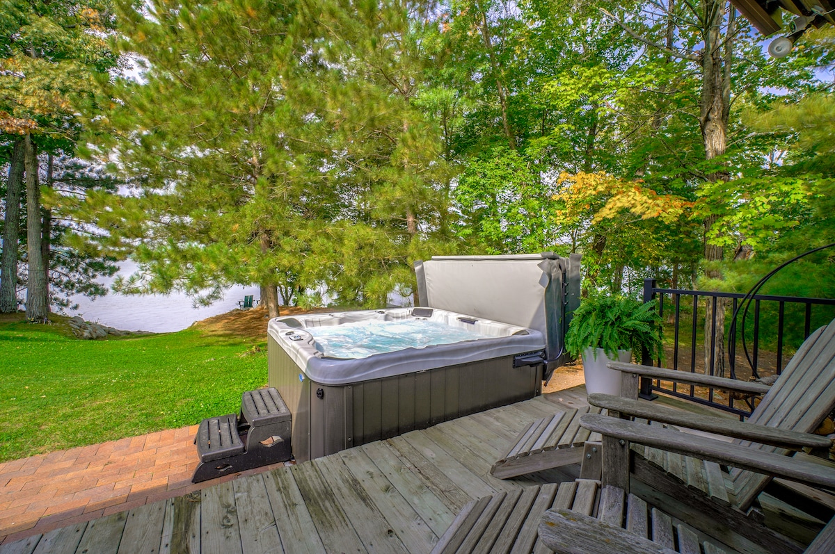 A private hot tub is situated on a wooden deck surrounded by lush green trees, offering a serene view of the lake. Adirondack chairs are placed nearby for relaxed seating. The natural setting enhances the tranquil atmosphere for unwinding outdoors.