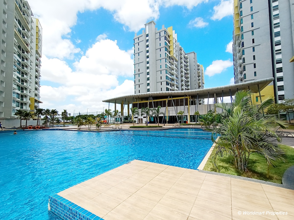 A large swimming pool is visible in the foreground, surrounded by a spacious deck and palm trees. Modern apartment buildings rise in the background under a clear blue sky dotted with clouds. An open-air pavilion is situated near the pool area, providing shaded seating.