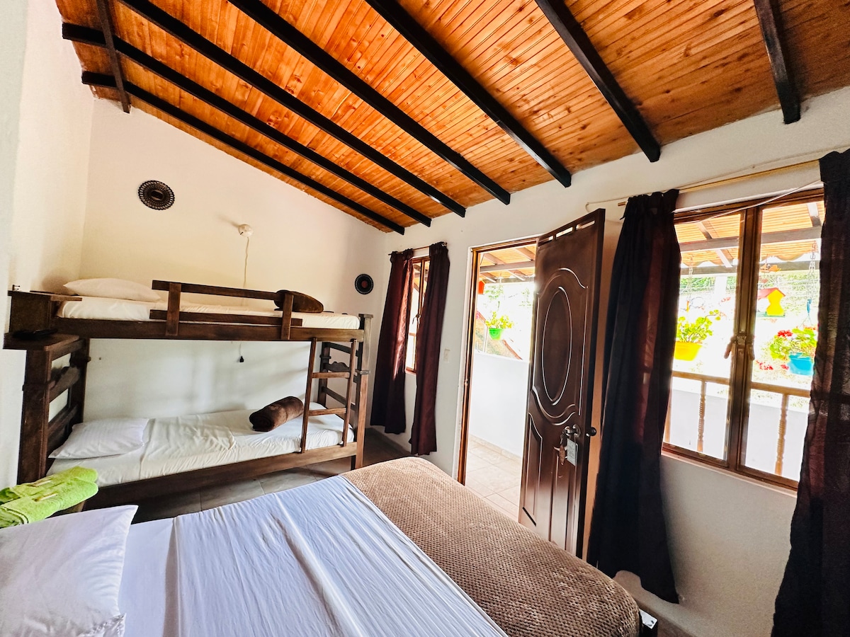 A well-lit sleeping area features a spacious bed alongside a sturdy wooden bunk bed. Light wood beams highlight the ceiling, while large windows provide a view of the outdoor space. Rich curtains frame the windows, complementing the neutral color scheme.
